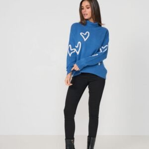A woman modeling a blue mock-neck knit sweater with white heart patterns, paired with black pants and black ankle boots, against a plain white studio background.