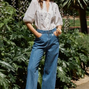 A woman stands outdoors in a garden setting wearing a white long-sleeve button-up blouse and high-waisted, wide-leg blue jeans with thin vertical pinstripes.