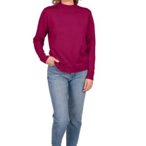 A woman modeling a long-sleeve mock neck sweater in deep magenta, paired with blue jeans and white sneakers, standing against a light gray studio background.